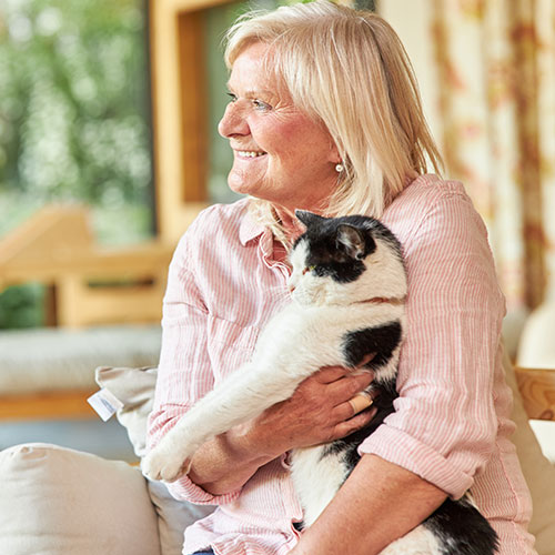 A smiling elderly woman with blonde hair, wearing a light pink shirt, is sitting and holding a black and white cat in her arms. The background shows a cozy home interior with large windows and blurred greenery outside, suggesting she might be reminiscing about her days as a veterinarian.