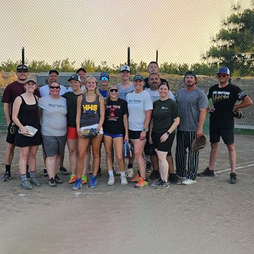 A group of people posing for a photo on a baseball field. The group consists of men and women of varying ages, including a veterinarian, dressed in casual sportswear and baseball caps. They stand closely together and smile at the camera, with a chain-link fence and trees in the background.