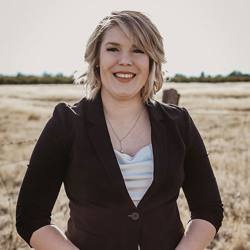 A woman with short blonde hair, wearing a dark blazer over a light top, stands smiling in a sunlit field of tall, dry grass with trees in the distant background.