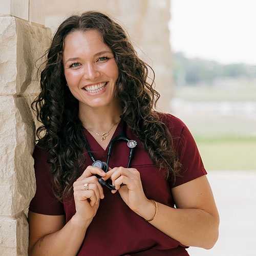 Woman with long curly hair, wearing a maroon shirt and stethoscope, smiles while leaning against a stone wall outdoors.